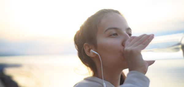 A young beautiful sportive girl listening to music and drinking water at sunrise by the lake.