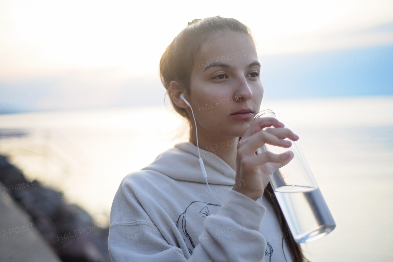 A young beautiful sportive girl listening to music and drinking water at sunrise by the lake.