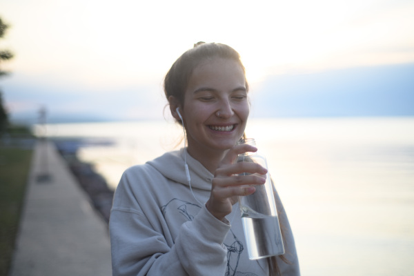 A young beautiful sportive girl listening to music and drinking water at sunrise by the lake.