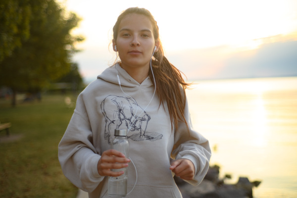 A young beautiful sportive girl listening to music at sunrise by the lake.