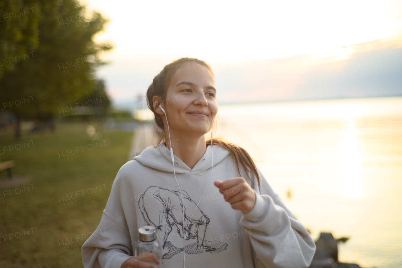 A young beautiful sportive girl listening to music and jogging at sunrise by the lake.