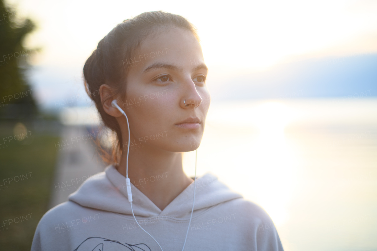 A young beautiful sportive girl listening to music at sunrise by the lake.