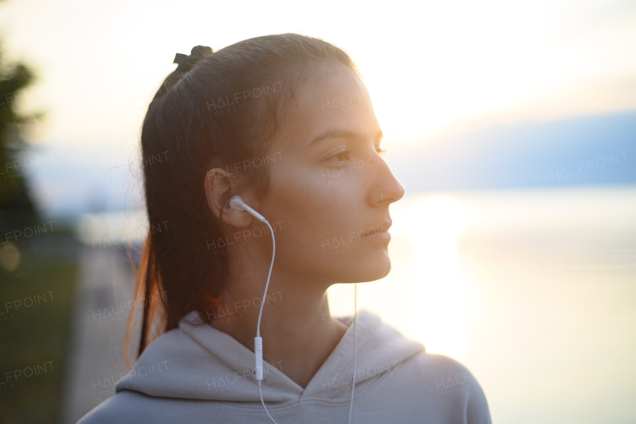 A young beautiful sportive girl listening to music at sunrise by the lake.