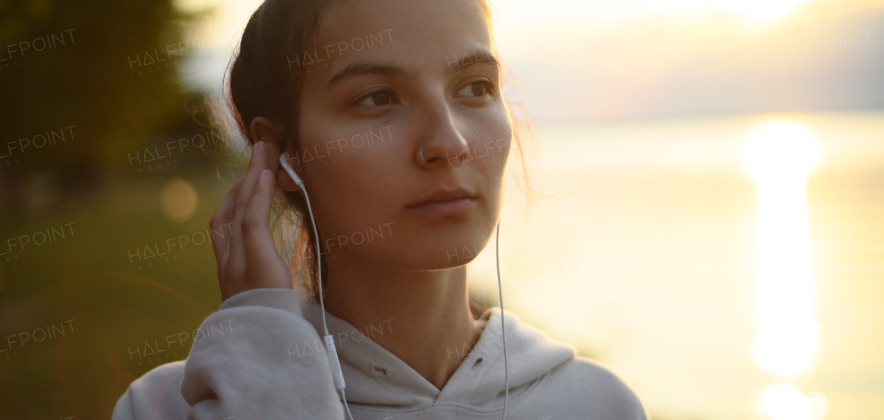 A young beautiful sportive girl listening to music at sunrise by the lake.