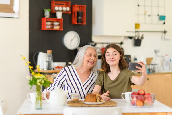 A happy senior grandmother with teenage granddaguhter sitting in kitchen and taking selfie with smartphone together at home.