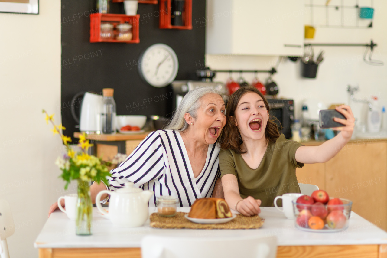 A happy senior grandmother with teenage granddaguhter sitting in kitchen and taking selfie with smartphone together at home.