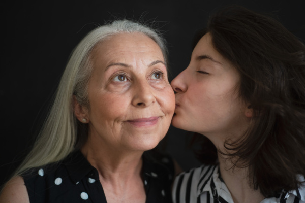 A portrait of senior grandmother with her granddaughter kissing her over black blackground