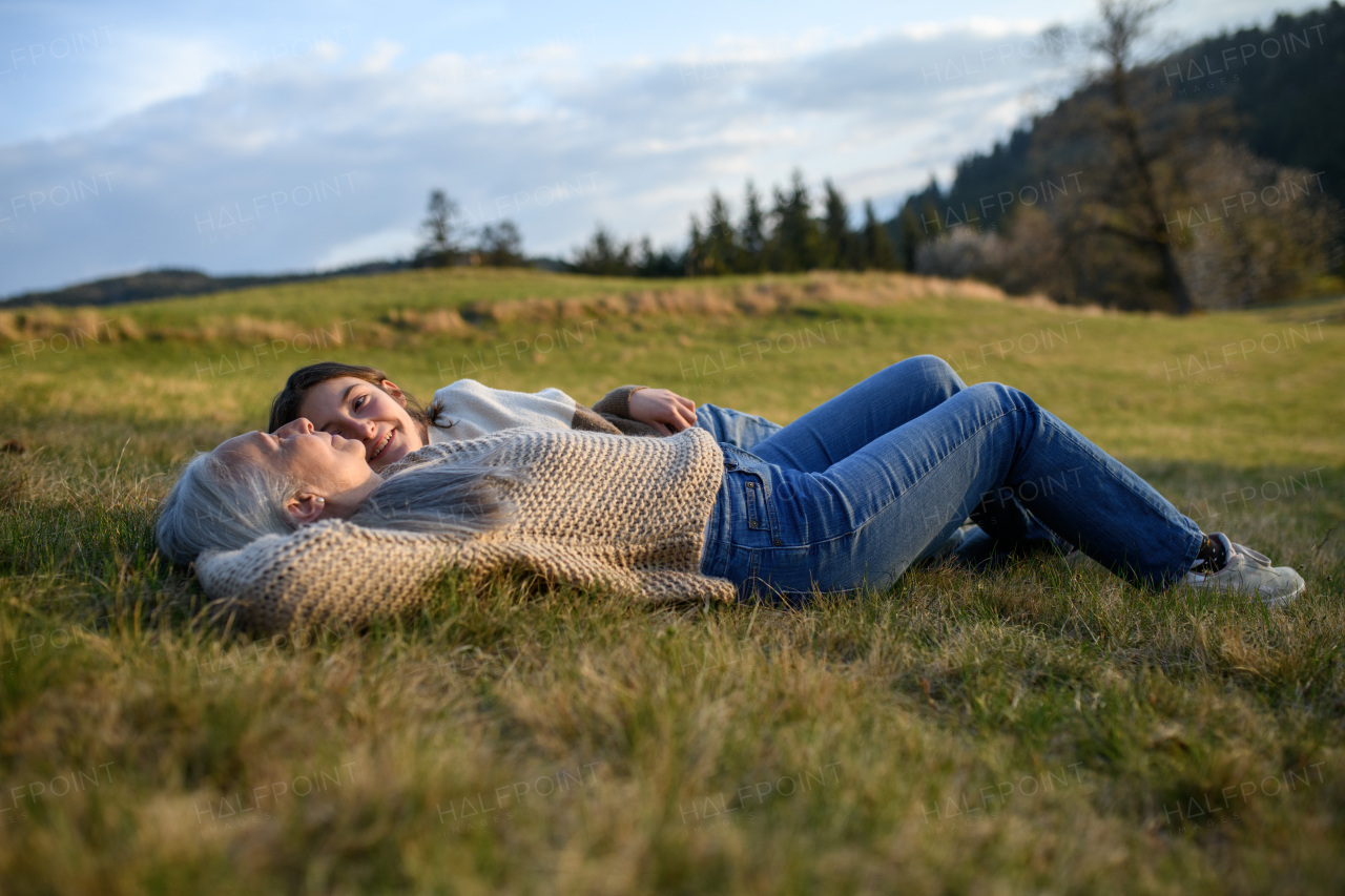 A happy senior grandmother with teenage granddaguhter lyinf down on grass in nature on spring day.