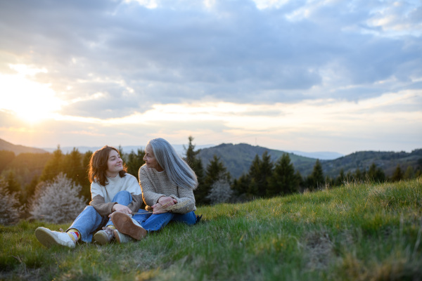 A happy senior grandmother with teenage granddaguhter sitting on grass in nature on spring day.