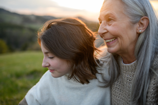 Portrait of a happy senior grandmother with teenage granddaguhter sitting on grass in nature on spring day during sunset.