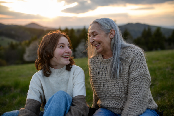 A happy senior grandmother with teenage granddaguhter sitting on grass in nature on spring day.