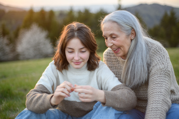 A happy senior grandmother with teenage granddaguhter sitting on grass and exploring nature on spring day.