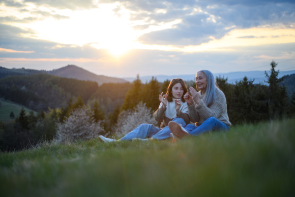 A happy senior grandmother with teenage granddaguhter sitting and whistling on grass in nature on spring day.