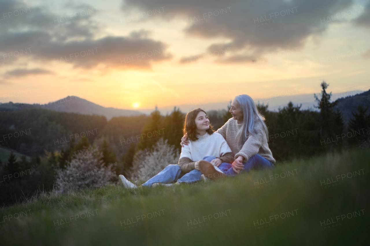 A happy senior grandmother with teenage granddaguhter sitting on grass in nature on spring day.