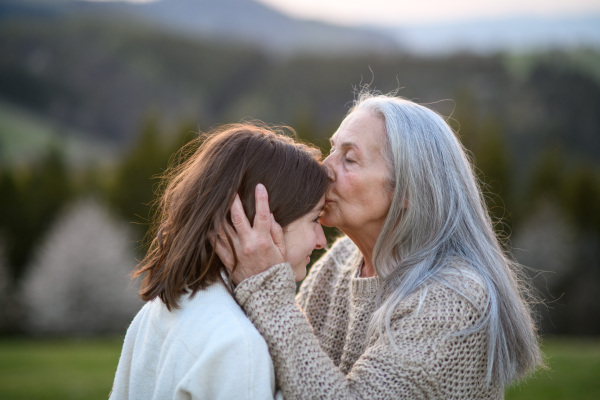 A happy senior grandmother kissing her teenage granddaguhter on forehead in nature on spring day.