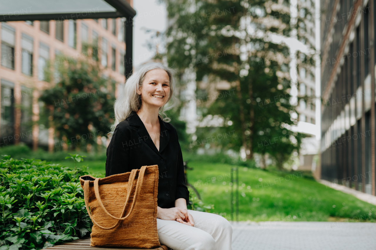 Portrait of beautiful mature woman in middle age with long gray hair, sitting on the bench in the city.