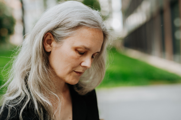 Portrait of pensive mature woman in middle age with long gray hair, standing outdoor in the city.