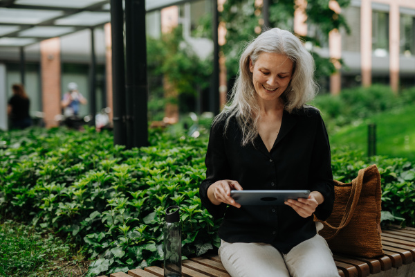 Portrait of beautiful mature woman in middle age with long gray hair, sitting on the bench in the city. Woman working outdoors, making videocall on tablet.