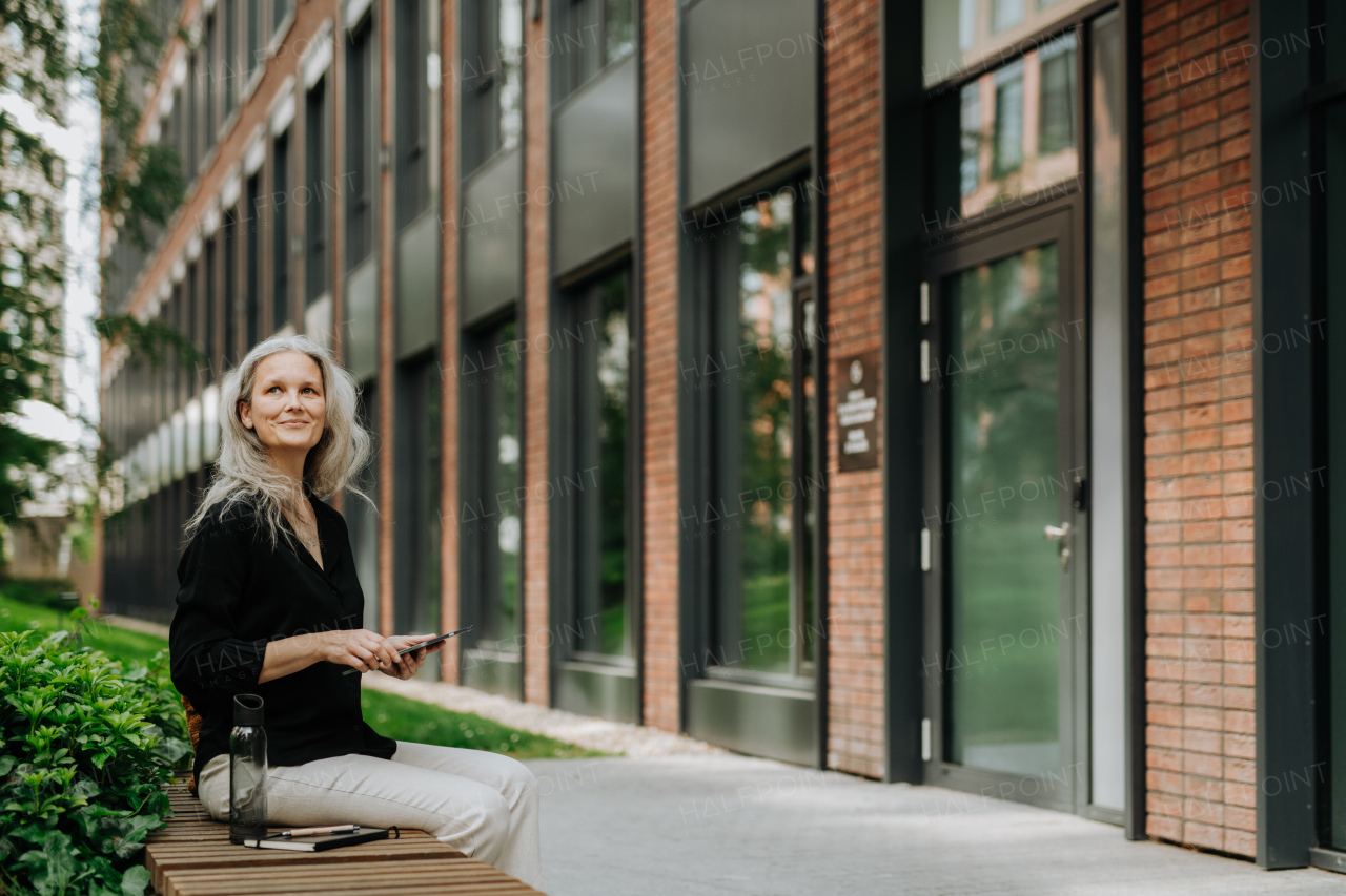 Portrait of beautiful mature woman in middle age with long gray hair, sitting on the bench in the city.