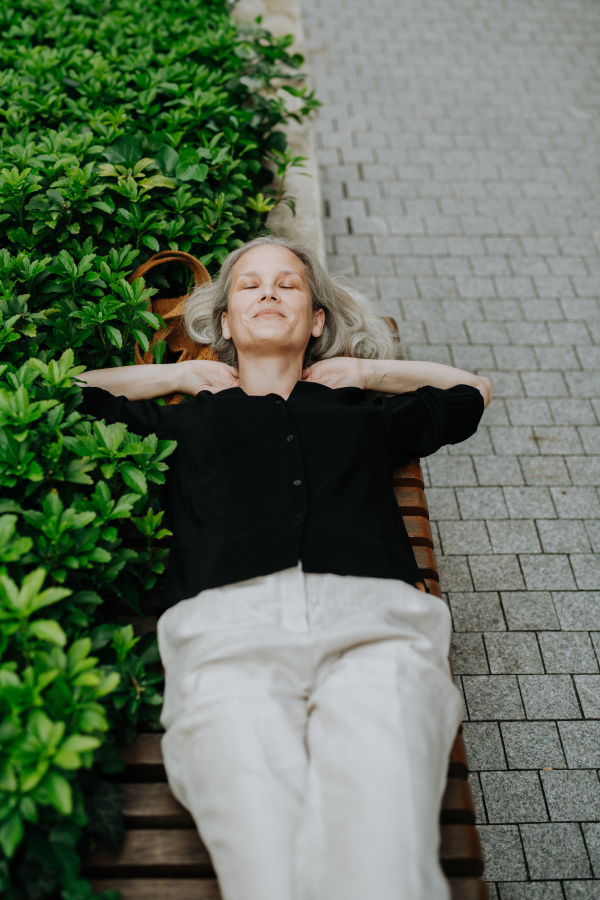 Shot from above, a beautiful mature woman in middle age with long gray hair, lying on the bench in city park. Smiling woman lying on her back outdoors, resting after workday. Banner with copy space.