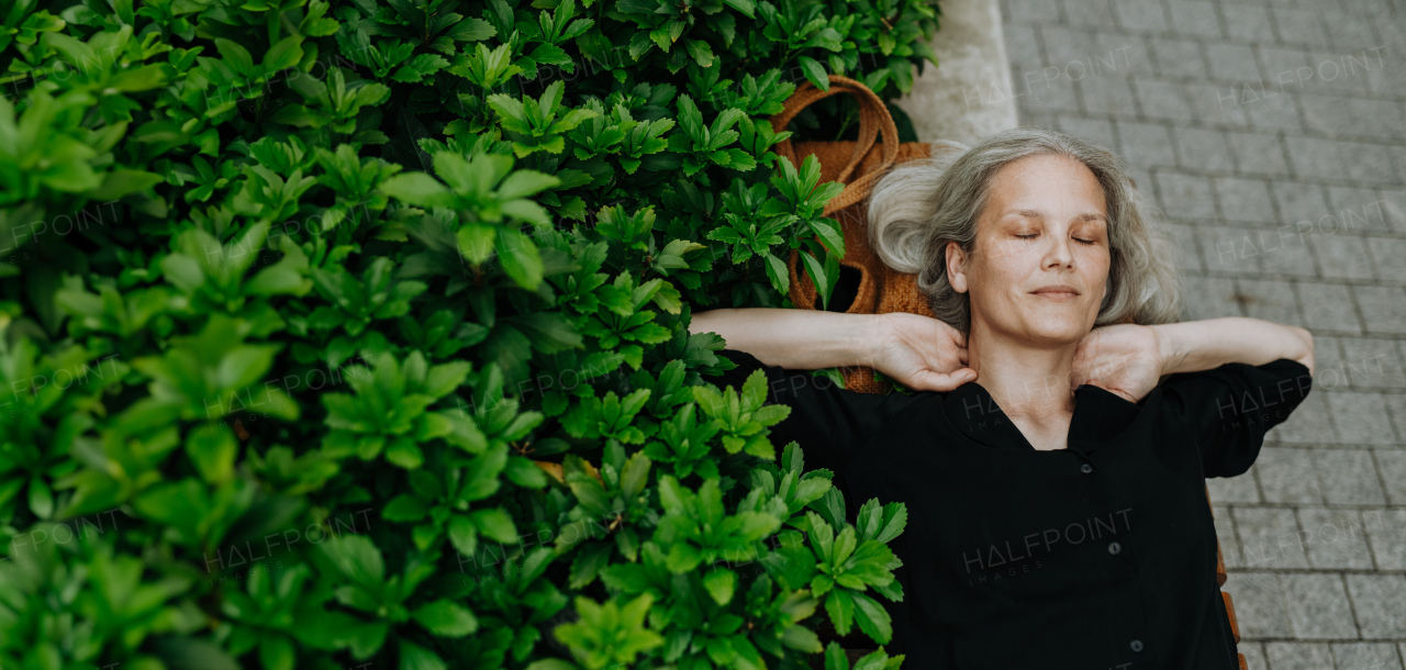 Shot from above, a beautiful mature woman in middle age with long gray hair, lying on the bench in city park. Smiling woman lying on her back outdoors, resting after workday. Banner with copy space.