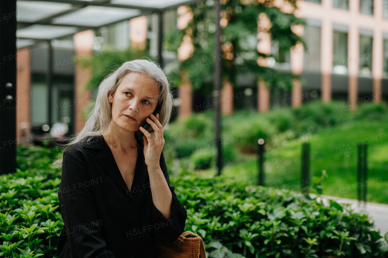 Portrait of beautiful mature woman in middle age with long gray hair, making a call outdoors in the city park.