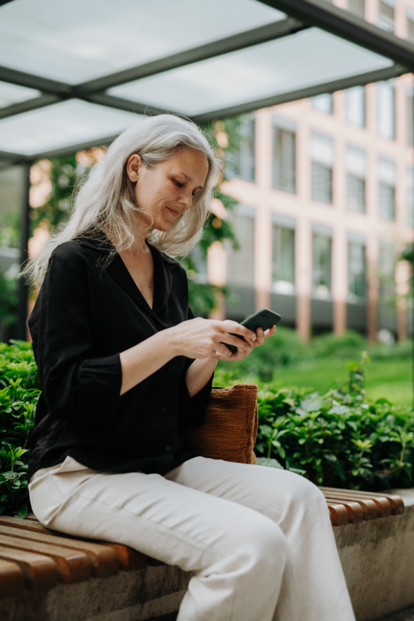 Portrait of beautiful mature woman in middle age with long gray hair, sitting on the bench in the city, holding smart phone.