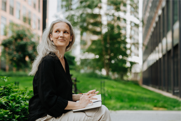 Portrait of beautiful mature woman in middle age with long gray hair, sitting on the bench in the city and writing in the notebook. Woman making checklist in her diary.