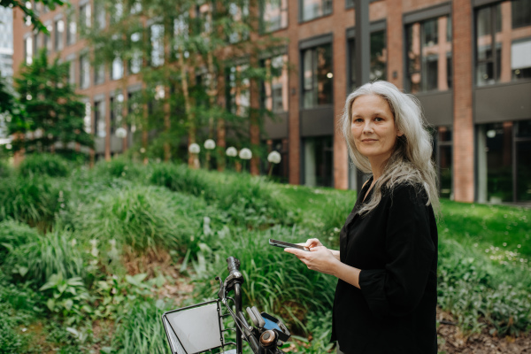 A beautiful middle-aged woman, who commutes through the city by bike, standing next to a bike rack and her bicycle. Female city commuter traveling from work by bike after a long workday. Renting city bike using mobile app.