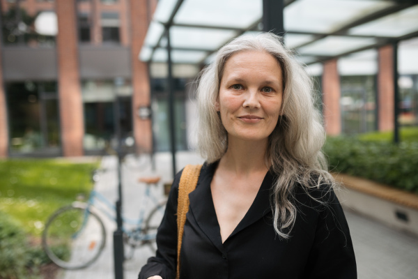 Portrait of middle-aged woman, who commutes through the city by bike, standing next to a bike rack and her bicycle. Female city commuter traveling from work by bike after a long workday. Renting city bike using mobile app.