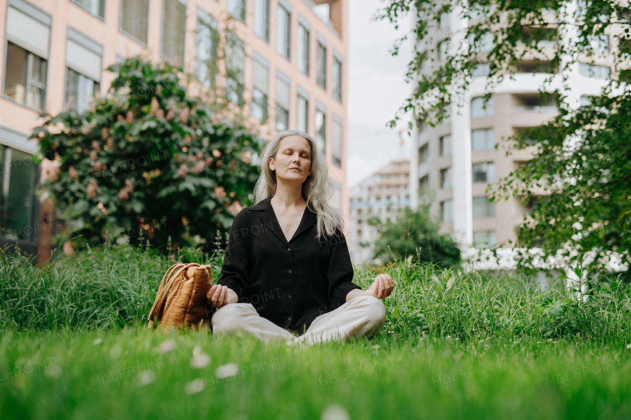 Portrait of beautiful mature woman in middle age with long gray hair, sitting on the grass in city park. Smiling woman meditating cross-legged on the grass, resting after workday.