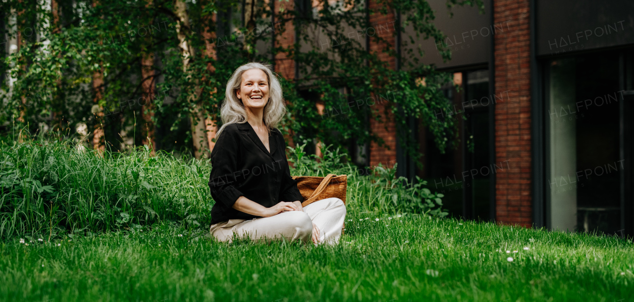 Portrait of beautiful mature woman in middle age with long gray hair, sitting on the grass in city park. Smiling woman sitting cross-legged on the grass, resting after workday. Banner with copy space.