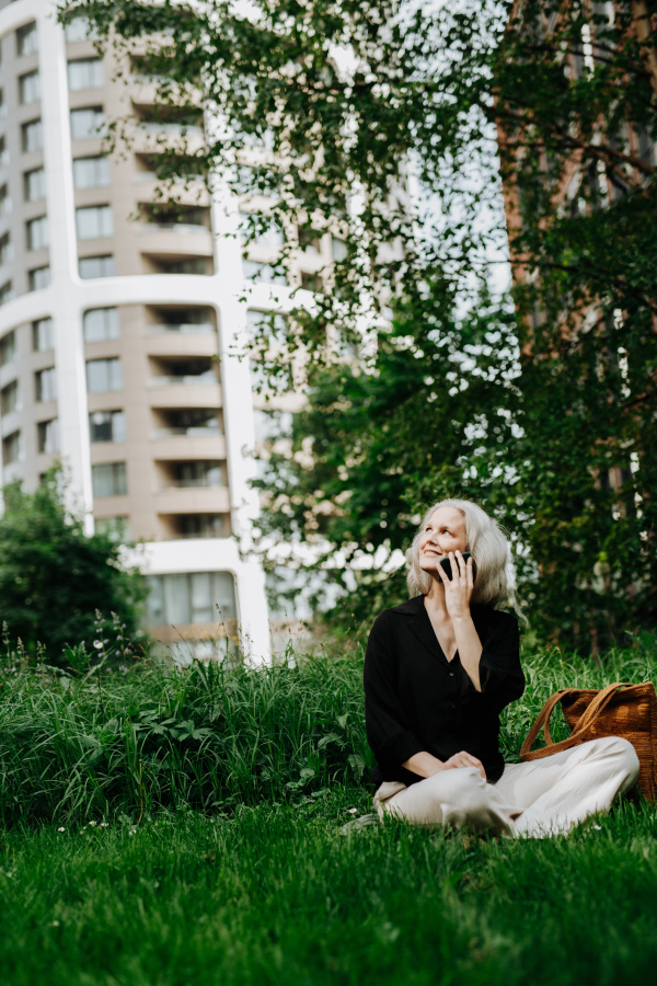 Portrait of beautiful mature woman in middle age with long gray hair, sitting on the grass in city park. Smiling woman sitting cross-legged on the grass, making a call.