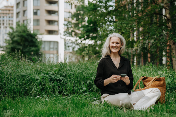 Portrait of beautiful mature woman in middle age with long gray hair, sitting on the grass in city park. Smiling woman sitting cross-legged on the grass, using her smartphone. Banner with copy space.