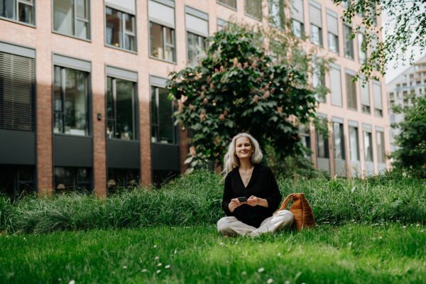 Portrait of beautiful mature woman in middle age with long gray hair, sitting on the grass in city park. Smiling woman sitting cross-legged on the grass, using her smartphone. Banner with copy space.