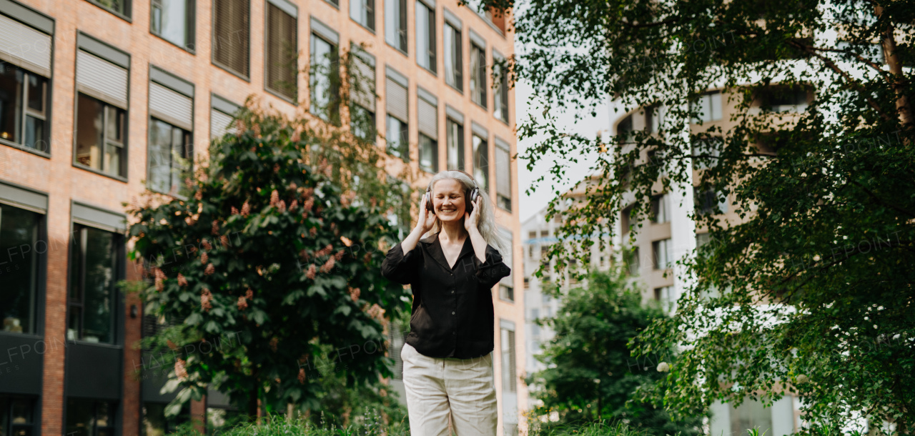 Portrait of a beautiful mature woman with gray hair, listening to music through wireless headphones in city park. Relaxing with music with closed eyes. Banner with copy space.