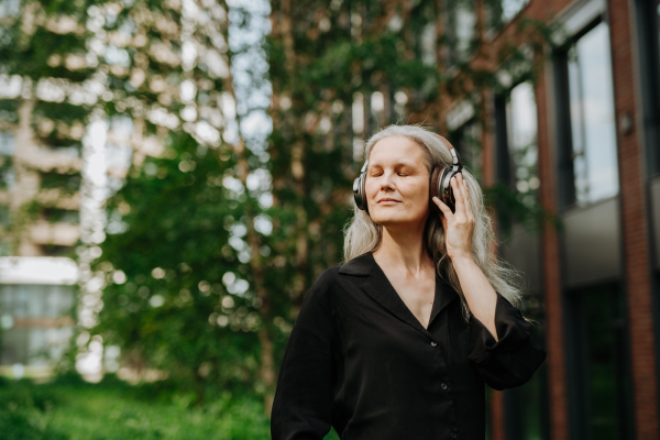 Portrait of a beautiful mature woman with gray hair, listening to music through wireless headphones in city park. Relaxing with music with closed eyes.