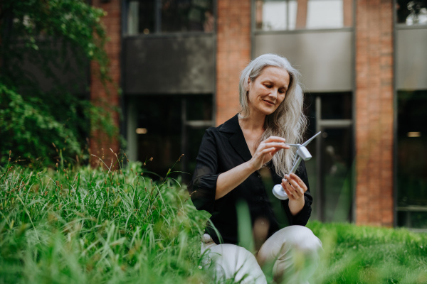 Portrait of beautiful mature woman in middle age with long gray hair, holding model of wind turbine. Concept of renewable resources. Importance of alternative energy sources and long-term sustainability.