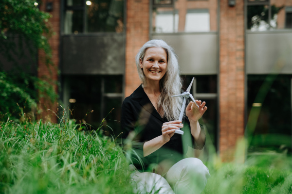 Portrait of beautiful mature woman in middle age with long gray hair, holding model of wind turbine. Concept of renewable resources. Importance of alternative energy sources and long-term sustainability.