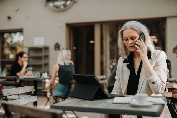 Portrait of a beautiful mature female manager with long gray hair, working outdoors in the city. The mature businesswoman working on tablet with keyboard while making a phone call on her smartphone.