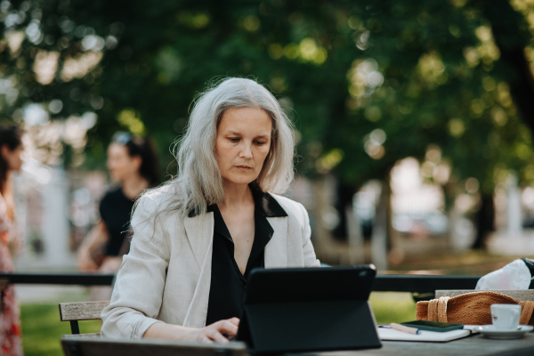 Portrait of a beautiful mature female manager with long gray hair, working outdoors in the city. The mature businesswoman is drinking coffee while having a video meeting on her tablet.