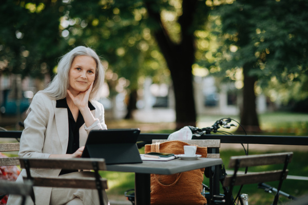 Portrait of a beautiful mature female manager with long gray hair, working outdoors in the city. The mature businesswoman is drinking coffee while having a video meeting on her tablet.