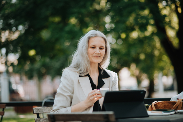 Portrait of a beautiful mature female manager with long gray hair, working outdoors in the city. The mature businesswoman is drinking coffee while having a video meeting on her tablet.