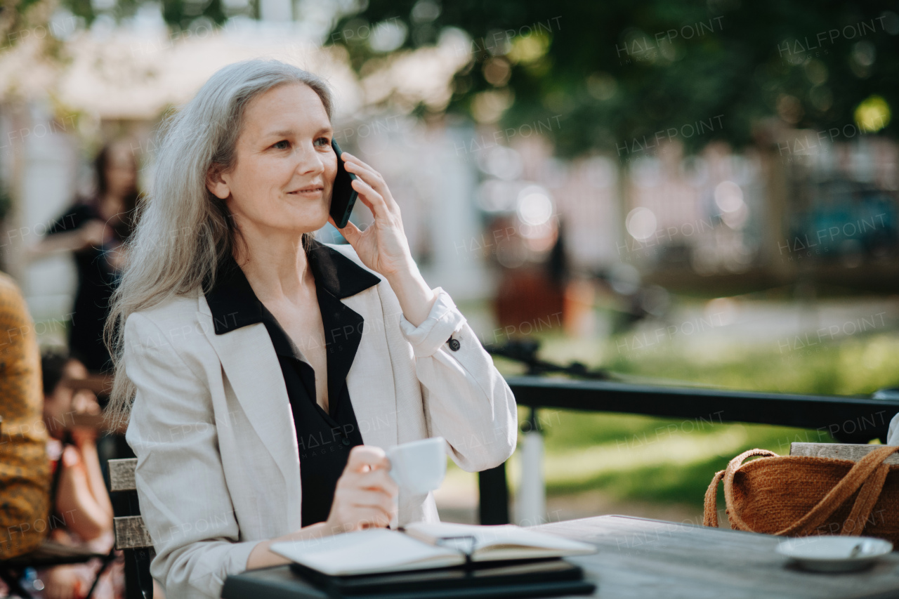 Portrait of a beautiful mature female manager with long gray hair, working outdoors in the city. The mature businesswoman is drinking coffee while making a phone call on her smartphone.