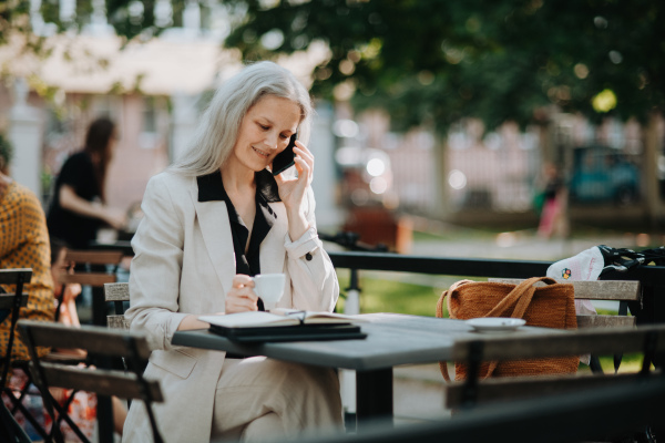 Portrait of a beautiful mature female manager with long gray hair, working outdoors in the city. The mature businesswoman is drinking coffee while making a phone call on her smartphone.