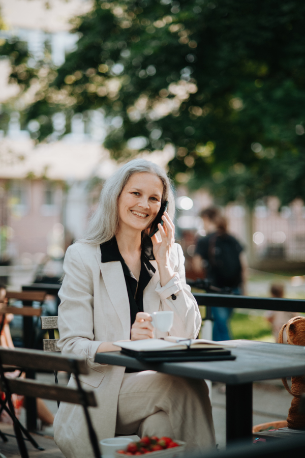 Portrait of a beautiful mature female manager with long gray hair, working outdoors in the city. The mature businesswoman is drinking coffee while making a phone call on her smartphone.