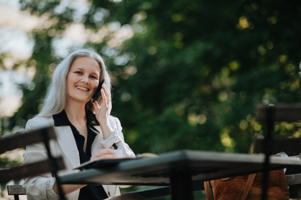 Portrait of a beautiful mature female manager with long gray hair, working outdoors in the city. The mature businesswoman is drinking coffee while making a phone call on her smartphone.