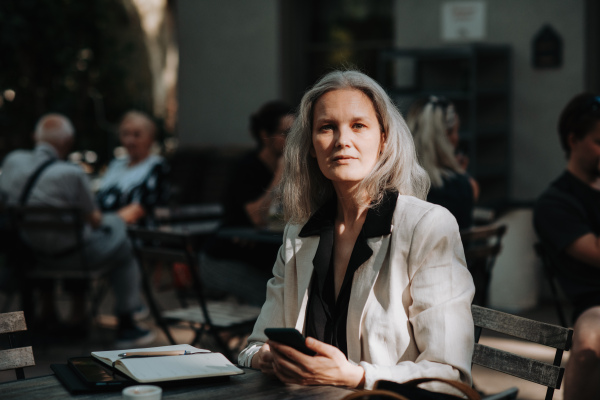 Portrait of a beautiful mature female manager with long gray hair, working outdoors in the city. The mature businesswoman is drinking coffee and writing in the diary. notepad.