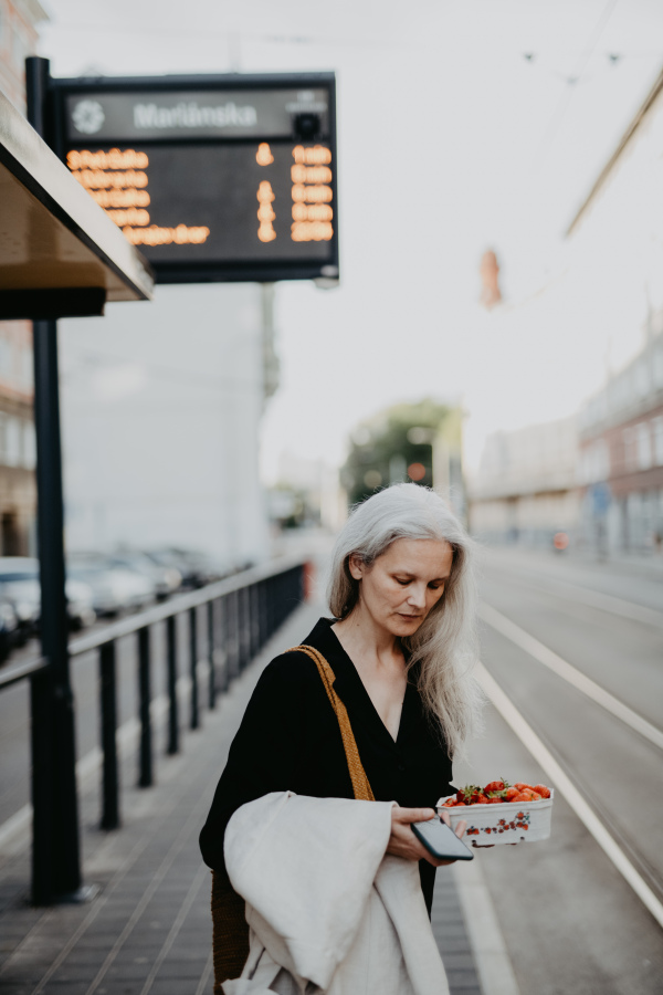 Portrait of a beautiful woman in middle age with gray hair, waiting for the bus at a bus stop. Female city commuter traveling from work by bus after a long workday.