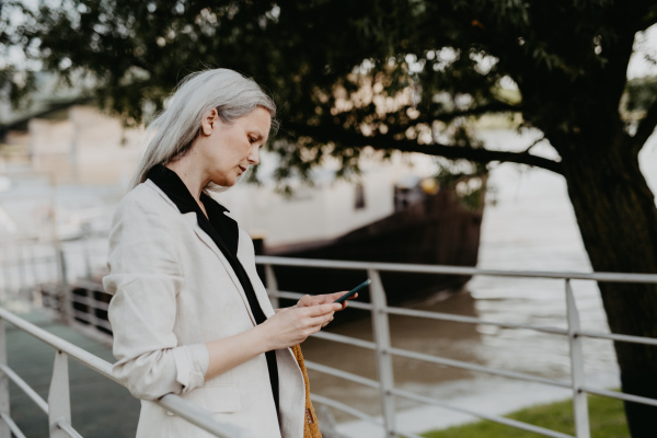 Portrait of beautiful mature woman in middle age with long gray hair, in city using her smartphone.